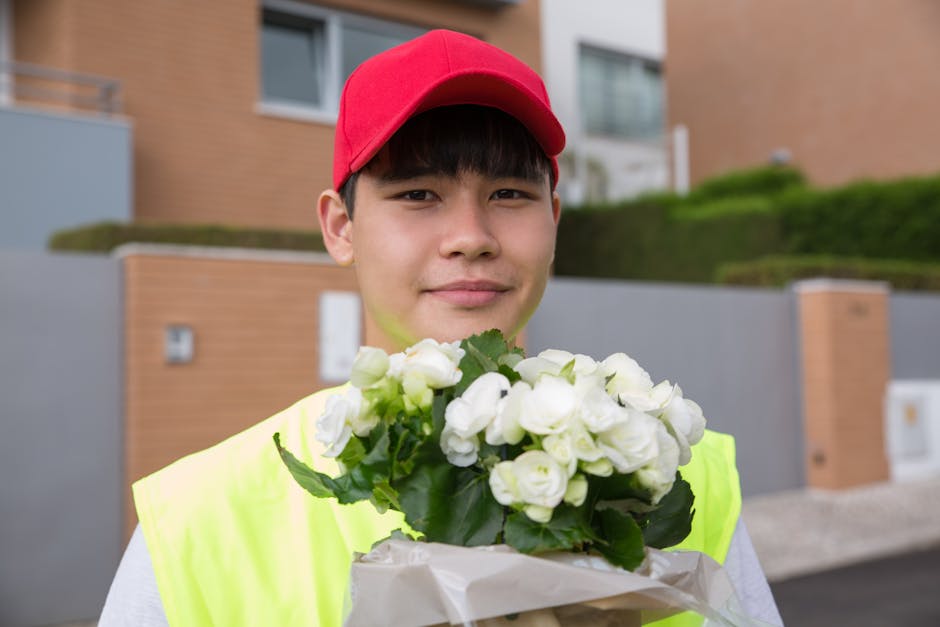 A young male delivery person wearing a red cap and a high-visibility yellow vest holds a bouquet of fresh white flowers, possibly roses and peonies, wrapped in white paper. He is standing outdoors in 