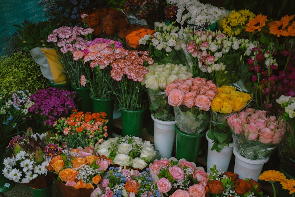 A vibrant display of fresh flower bouquets and potted arrangements arranged in green and white vases and wrapping, featuring a variety of flowers such as pink roses, white roses, yellow roses, orange 